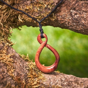 May include: A wooden pendant in the shape of an infinity symbol, carved from reddish-brown wood, hanging from a black cord against a mossy tree branch background.
