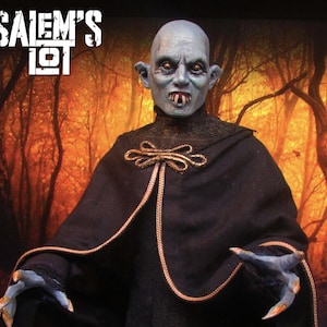 May include: A detailed figure of a vampire with pale blue skin, pointed ears, and sharp fangs. The figure wears a black cloak with gold accents and has long, slender fingers. The text "Salem's Lot" is displayed in white at the top.