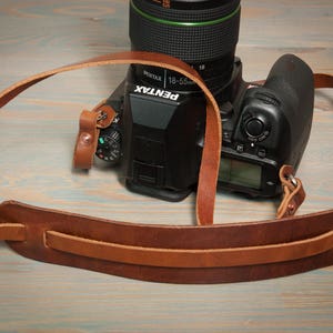 May include: A brown leather camera strap with a padded shoulder section is draped over a black Pentax DSLR camera. The strap is made of two shades of brown leather and is attached to the camera body. The lens is black with a green accent.