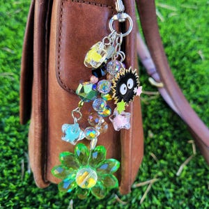 May include: A brown leather handbag with a decorative charm. The charm features a green flower, colourful beads, and a black character with white eyes. The background is green grass.