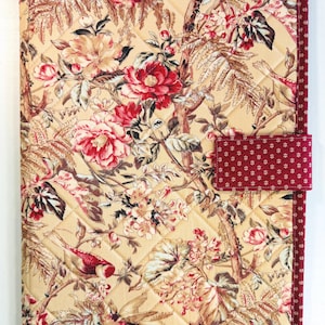 May include: A rectangular quilted fabric cover featuring a cream background with a floral and bird pattern in shades of red, brown, and green. The cover is edged with a dark red fabric border with a small white geometric print.