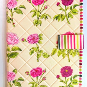 May include: A quilted fabric book cover featuring a cream background with a repeating pattern of pink and magenta roses and green leaves. The edges and the horizontal closure strap are finished with a multi-colored vertical striped fabric trim.