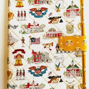 May include: A quilted fabric cover featuring a repeating pattern of American historical icons including the Liberty Bell, eagles, cannons, drums, colonial buildings, and scrolls labeled In Congress July 4 1776, finished with a mustard yellow border and tab.