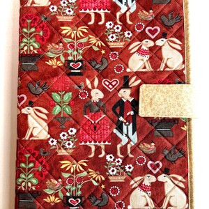 May include: A rectangular quilted fabric cover featuring a dark red background with a repeating pattern of rabbits in formal attire, potted flowers, birds, and hearts. The cover has a beige floral-patterned border and a matching side tab closure.