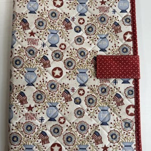 May include: A quilted fabric book cover featuring a cream background with a repeating pattern of blue vases labeled 1776, American flags, red stars, and the text BRIGHT STARS. The cover has a red fabric border and a matching red fabric closure tab.
