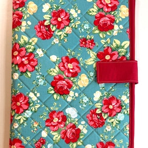 May include: A rectangular quilted fabric cover featuring a teal background with a repeating pattern of red roses, green leaves, and small yellow flowers. The cover is edged with solid red fabric binding and includes a matching red fabric tab closure.