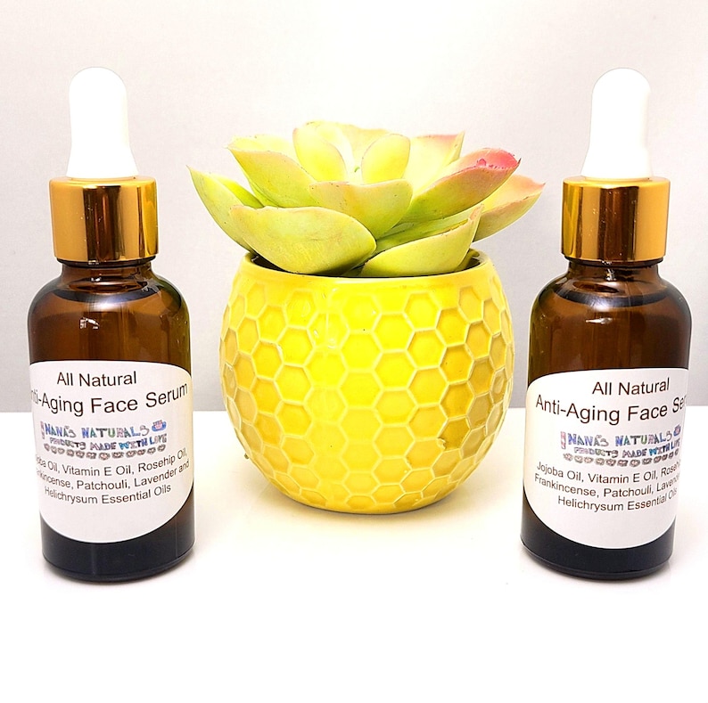 May include: Two amber glass bottles with gold lids and droppers filled with a clear liquid. The bottles are labeled "All Natural Anti-Aging Face Serum" and contain ingredients like jojoba oil, vitamin E oil, rosehip oil, frankincense, patchouli, lavender, and helichrysum essential oils. The bottles are sitting on a white surface in front of a yellow planter with a green succulent plant.