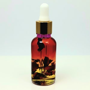 May include: A clear glass bottle with a gold lid and a white dropper. The bottle is filled with a reddish-brown liquid and contains dried rose petals.