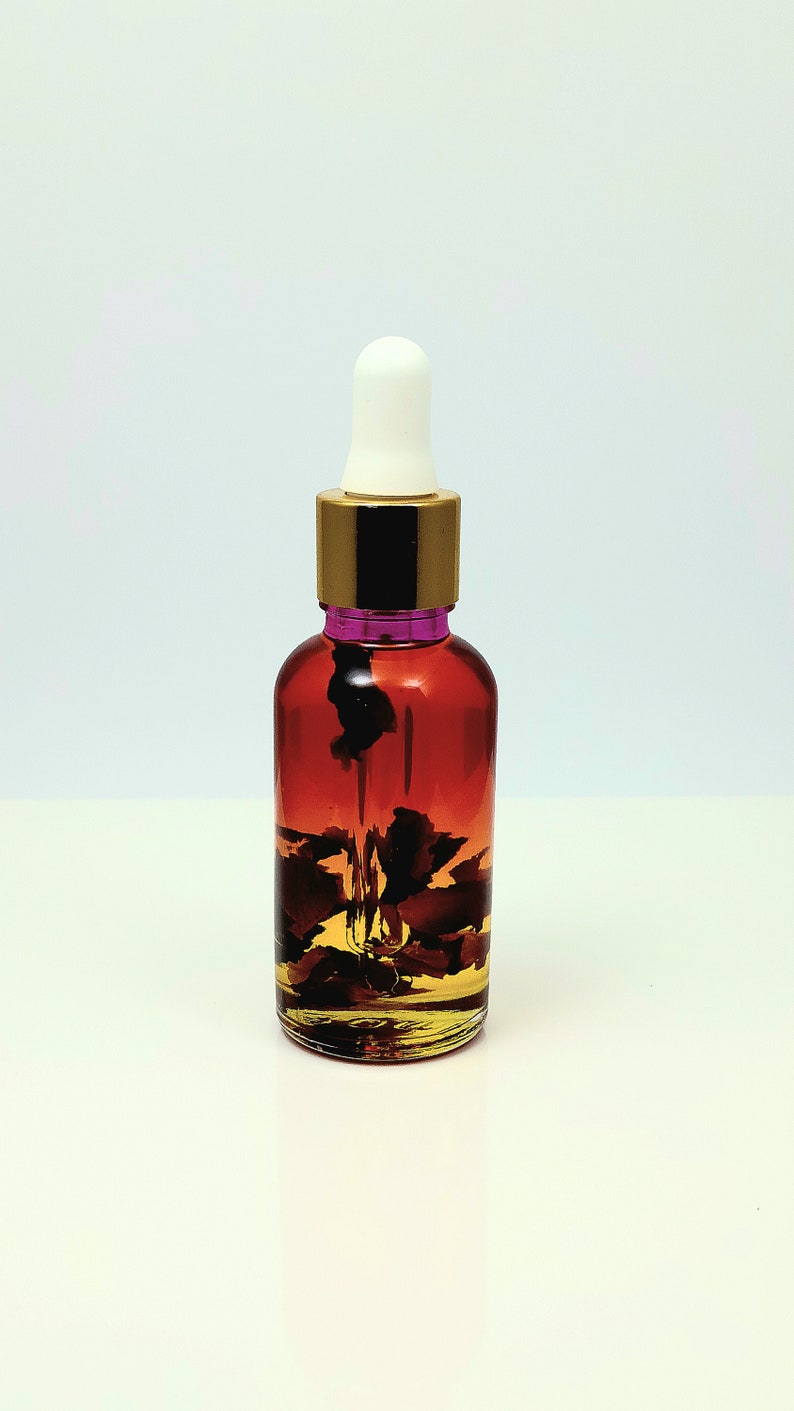 May include: A clear glass bottle with a gold lid and a white dropper. The bottle is filled with a reddish-brown liquid and contains dried rose petals.