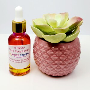 May include: A clear glass bottle with a gold lid and a white dropper top. The bottle is filled with a yellow liquid and has a pink label that reads "All Natural Rose Face Serum Nana's Naturals Products Made with Love Jojoba Oil, Vitamin E Oil, Rosehip Oil, Rose and Jasmine Essential Oils". The bottle is sitting on a white surface next to a pink ceramic pot with a green succulent plant.