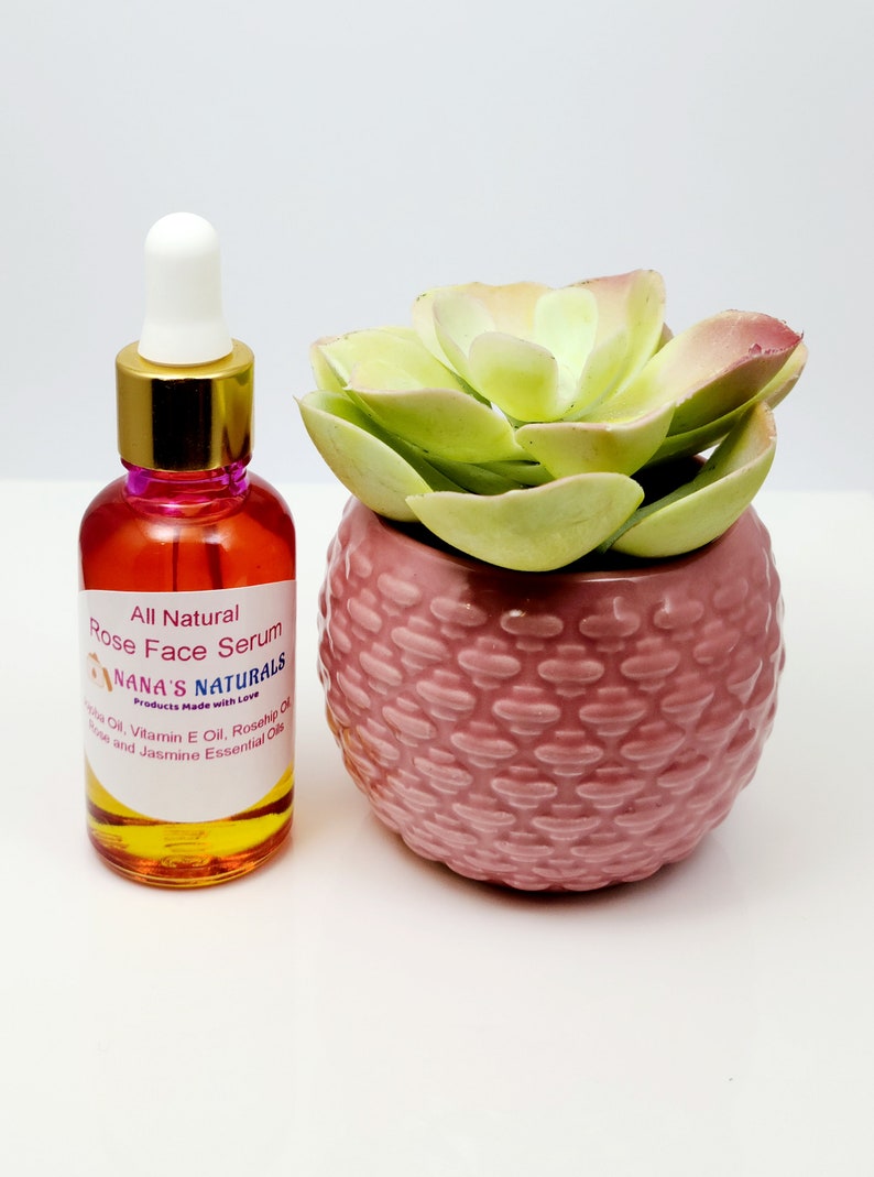 May include: A clear glass bottle with a gold lid and a white dropper top. The bottle is filled with a yellow liquid and has a pink label that reads "All Natural Rose Face Serum Nana's Naturals Products Made with Love Jojoba Oil, Vitamin E Oil, Rosehip Oil, Rose and Jasmine Essential Oils". The bottle is sitting on a white surface next to a pink ceramic pot with a green succulent plant.