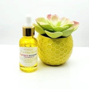 May include: A glass bottle of all-natural citrus face serum with a gold lid and dropper. The bottle is sitting on a white surface next to a yellow planter with a green succulent.