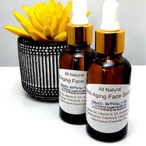 May include: Two amber glass bottles with gold lids and white droppers filled with a brown liquid. The bottles are labeled "All Natural Anti-Aging Face Serum" and feature the text "NANA'S NATURALS MADE WITH LOVE". The bottles are sitting on a white surface.