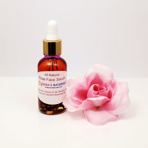 May include: A small amber glass bottle with a gold lid and a white dropper. The bottle contains a rose face serum. The label reads "All Natural Rose Face Serum Nana's Naturals Products Made with Love Jojoba Oil, Vitamin E Oil, Rosehip Oil, Rose and Jasmine Essential Oils". A pink rose is in the background.