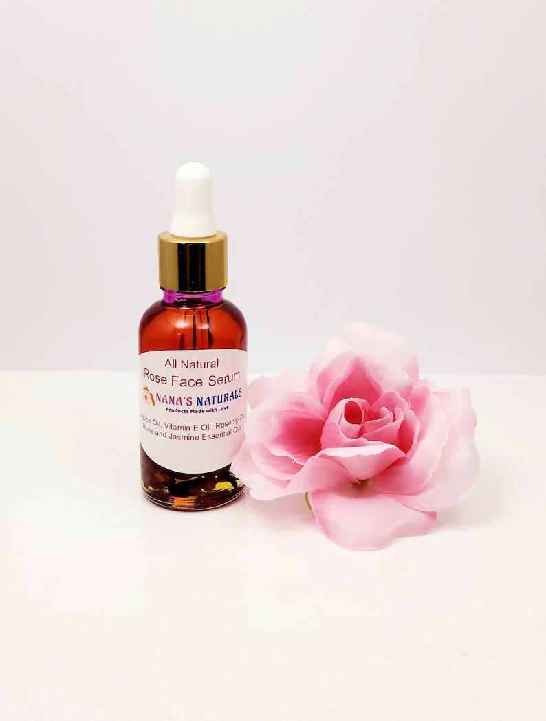 May include: A small amber glass bottle with a gold lid and a white dropper. The bottle contains a rose face serum. The label reads "All Natural Rose Face Serum Nana's Naturals Products Made with Love Jojoba Oil, Vitamin E Oil, Rosehip Oil, Rose and Jasmine Essential Oils". A pink rose is in the background.