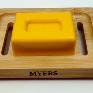 May include: A yellow bar of soap sits on a wooden soap dish with the word "MYERS" printed on the front.
