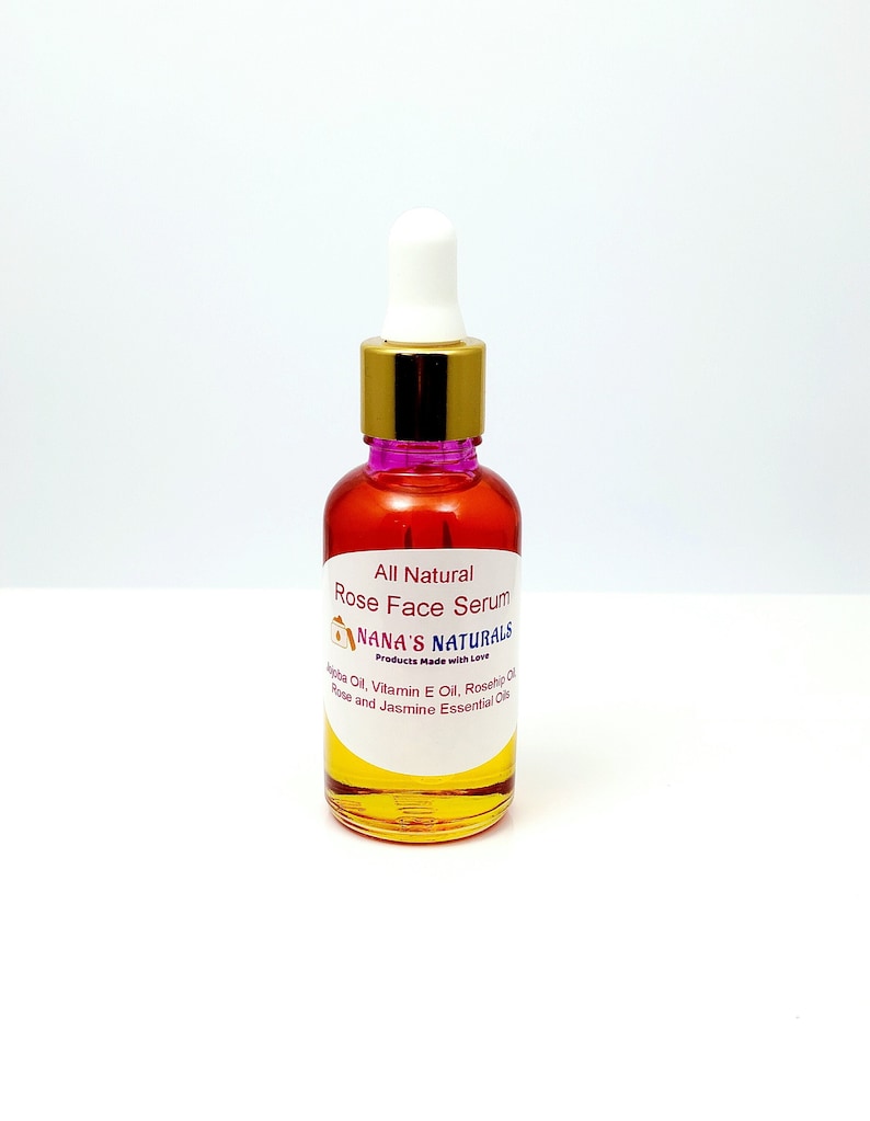 May include: A clear glass bottle with a gold lid and a pink dropper. The bottle contains a yellow liquid and has a white label with the text "All Natural Rose Face Serum Nana's Naturals Products Made with Love Jojoba Oil, Vitamin E Oil, Rosehip Oil, Rose and Jasmine Essential Oils".