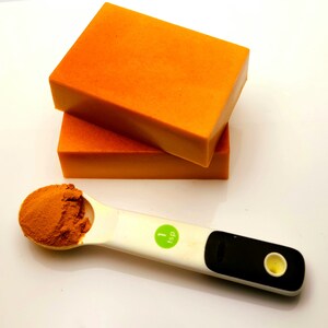 May include: Two orange bars of soap stacked on top of each other. A white measuring spoon with a green label that says "1 tsp" is filled with orange powder.