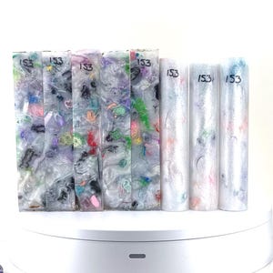 May include: Eight clear resin pen blanks with embedded colorful flecks and swirls. The blanks are labeled "153" and are arranged in a row on a white surface.