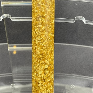 May include: A gold and clear resin pen blank with a speckled pattern. The blank is cylindrical and has a smooth surface.