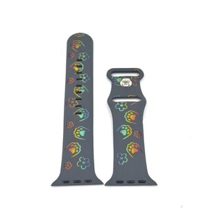 May include: Dark gray Apple Watch band featuring a repeating pattern of colorful paw prints and flowers. The band is made of a flexible material and has a buckle closure. The design includes rainbow-colored accents.