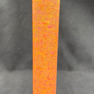 May include: A clear cylinder filled with a translucent orange liquid with pink and yellow glitter suspended inside.