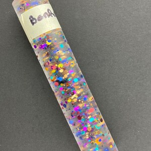 Może przedstawiać: Przezroczysty cylinder wypełniony kolorowym brokatem, w tym złotym, niebieskim, fioletowym i czerwonym. Cylinder ma białą etykietę z napisem "Bangle" napisanym czarnym markerem.