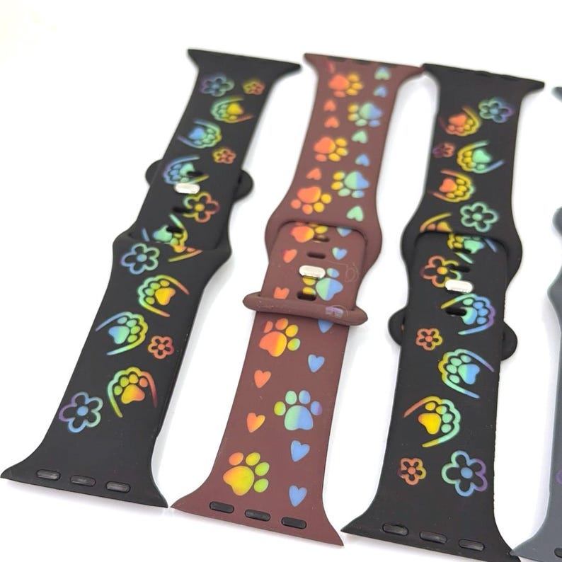 May include: A collection of watch bands in various colors, featuring paw prints, flowers, and hearts. The bands are black and brown, with rainbow-colored designs. The watch bands are made of a flexible material.