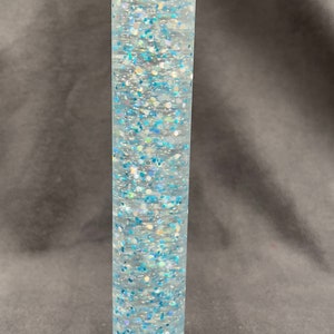 May include: A clear glass cylinder filled with blue and iridescent glitter. The glitter is suspended in a clear liquid and moves around when the cylinder is tilted.