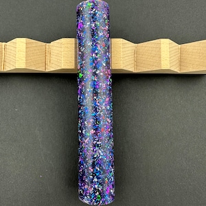May include: A dark blue pen blank with a variety of colorful glitter embedded in the resin. The blank is cylindrical and has a smooth, polished finish.