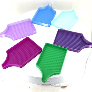May include: A collection of seven colorful plastic trays in shades of purple, blue, and green. Each tray has a rectangular base with vertical grooves and a curved handle. The trays are arranged in a circular pattern.