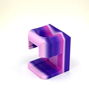 May include: A 3D printed phone stand in a purple and pink ombre color scheme. The stand is designed to hold a phone upright.
