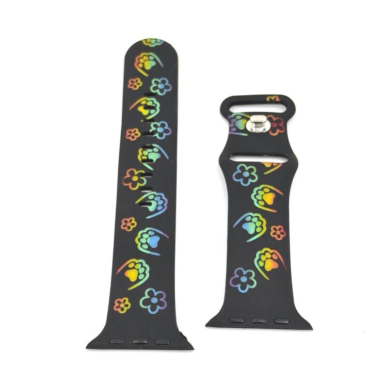 May include: Black watch band with a repeating pattern of colorful paw prints and flowers. The paw prints and flowers are in a rainbow gradient. The band is made of a flexible material and has a buckle closure.