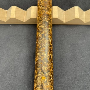 May include: A long, cylindrical, clear resin pen blank with gold flakes embedded throughout. The blank is approximately 10 inches long and 1 inch in diameter.