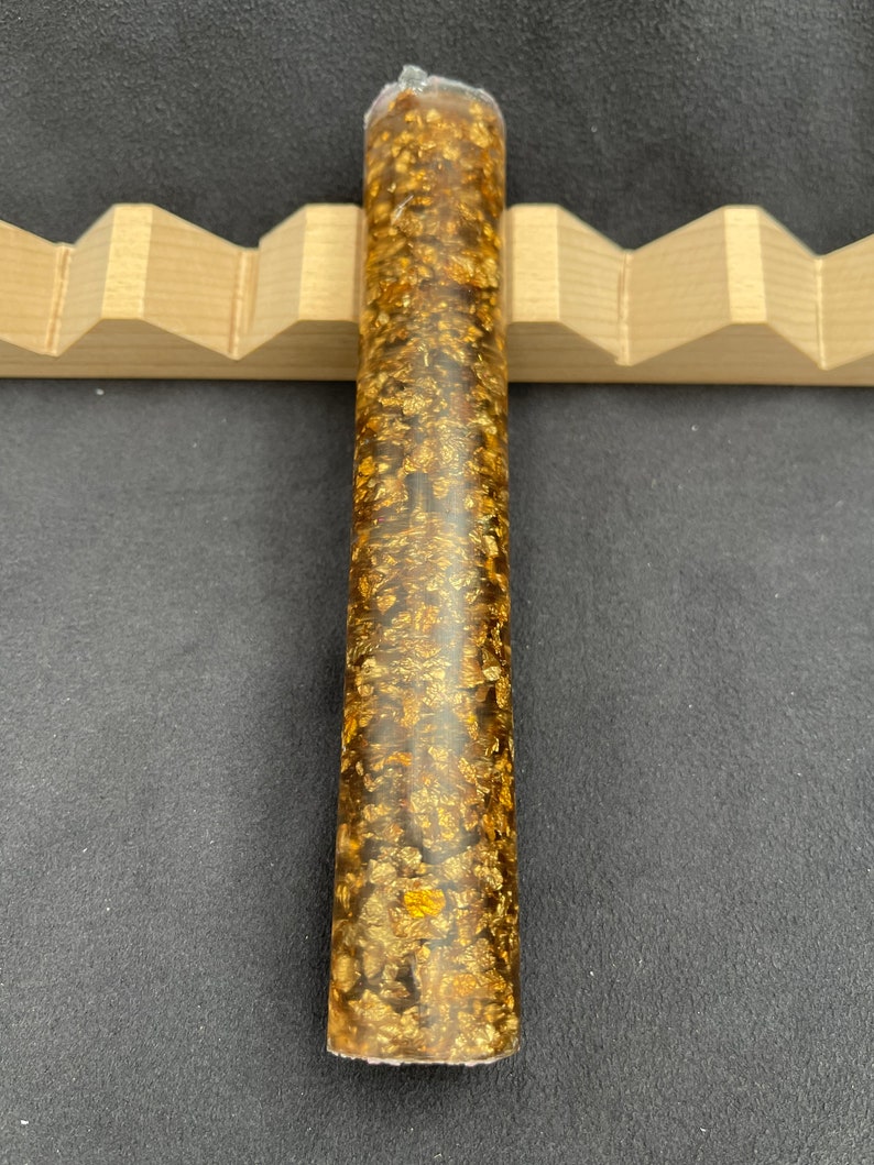 May include: A long, cylindrical, clear resin pen blank with gold flakes embedded throughout. The blank is approximately 10 inches long and 1 inch in diameter.