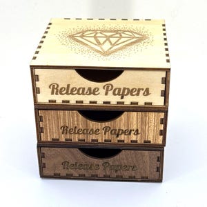 May include: A three-tiered wooden box with drawers. The top drawer is light-colored with a diamond design. The middle drawer is a medium brown, and the bottom drawer is dark brown. Each drawer is labeled "Release Papers".