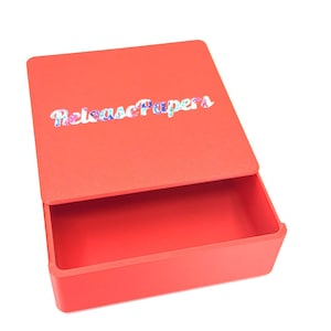May include: A rectangular red storage box with a sliding lid. The top of the lid features the word ReleasePapers printed in a multi-colored, floral-patterned font. The box is open, revealing a hollow interior space for storage and organization.