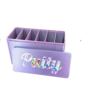 May include: A rectangular lavender storage container with six internal compartments and a matching lid. The lid features the word Putty in a colourful, multi-patterned, mosaic-style font. The container has a textured, glittery finish throughout.