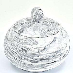 May include: A round, lidded container with a marbled design in shades of gray and white. The lid features a small, spherical handle. The container's surface has a swirling pattern, creating a unique, decorative appearance. The item is likely a decorative storage jar or a similar household item.