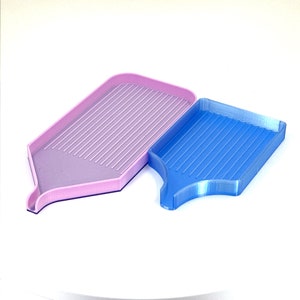 May include: Two plastic diamond painting trays. One is light pink, the other is blue. Both trays have a rectangular shape with a sloped bottom and a spout. The trays have parallel grooves for organizing diamond painting drills.