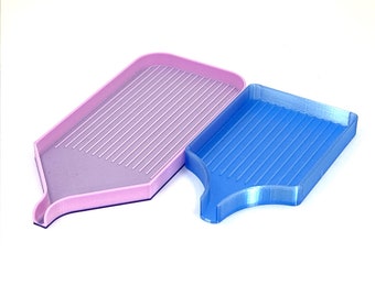 May include: Two plastic diamond painting trays. One is light pink, the other is blue. Both trays have a rectangular shape with a sloped bottom and a spout. The trays have parallel grooves for organizing diamond painting drills.