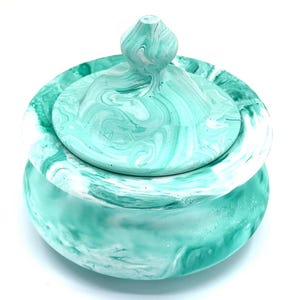 May include: A round, lidded container with a swirling teal and white pattern. The container has a rounded base and a matching lid with a small, decorative knob. The marbled effect gives the piece a unique, artistic look. The container is likely made of resin or a similar material.