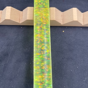 May include: A green and orange resin pen blank with iridescent glitter. The blank is cylindrical and has a smooth surface.