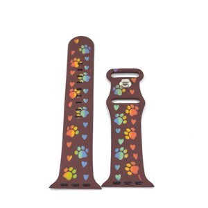 May include: A brown watch band with a pattern of rainbow paw prints and hearts. The band is made of a flexible material and has a metal clasp. The design features a repeating pattern of colorful paw prints and small hearts.