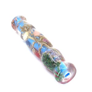 May include: A cylindrical resin object with a smooth, clear finish, containing embedded dried flowers, green foliage, and wheat stalks. The interior features a vibrant blue core, with the overall structure showing a slightly wavy, textured surface.