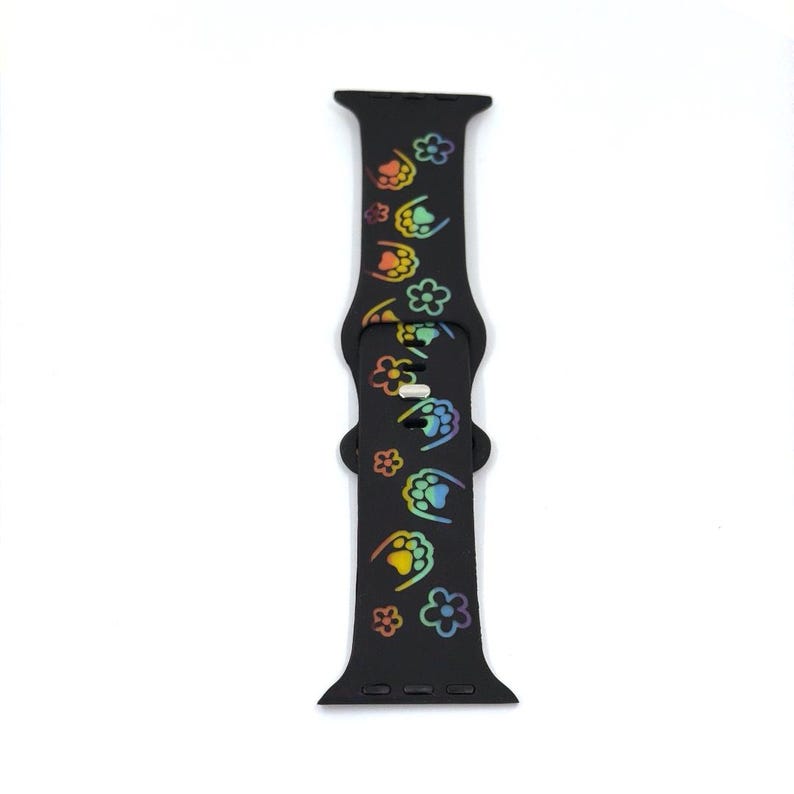 May include: Black watch band with a repeating pattern of colorful paw prints and flowers. The band is made of a flexible material and has a buckle closure. The paw prints and flowers are in a rainbow of colors.