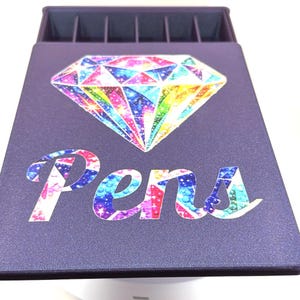 May include: A dark purple rectangular storage case with a top compartment featuring six slots. The lid displays a large, colourful, multi-faceted diamond graphic above the word Pens, which is written in a stylised, rainbow-coloured script font.