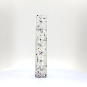 May include: A clear glass tube filled with white and purple glitter. The glitter is iridescent and creates a shimmering effect.
