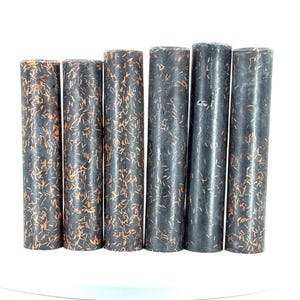 May include: Six cylindrical objects with a dark, speckled appearance. The cylinders are black with copper-colored flecks throughout. They are arranged vertically against a white background. The objects are of varying heights.