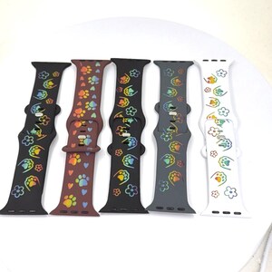 May include: Five colorful watch bands with paw prints and flower designs. The bands are in black, brown, black, gray, and white. The paw prints and flowers have a rainbow gradient effect.